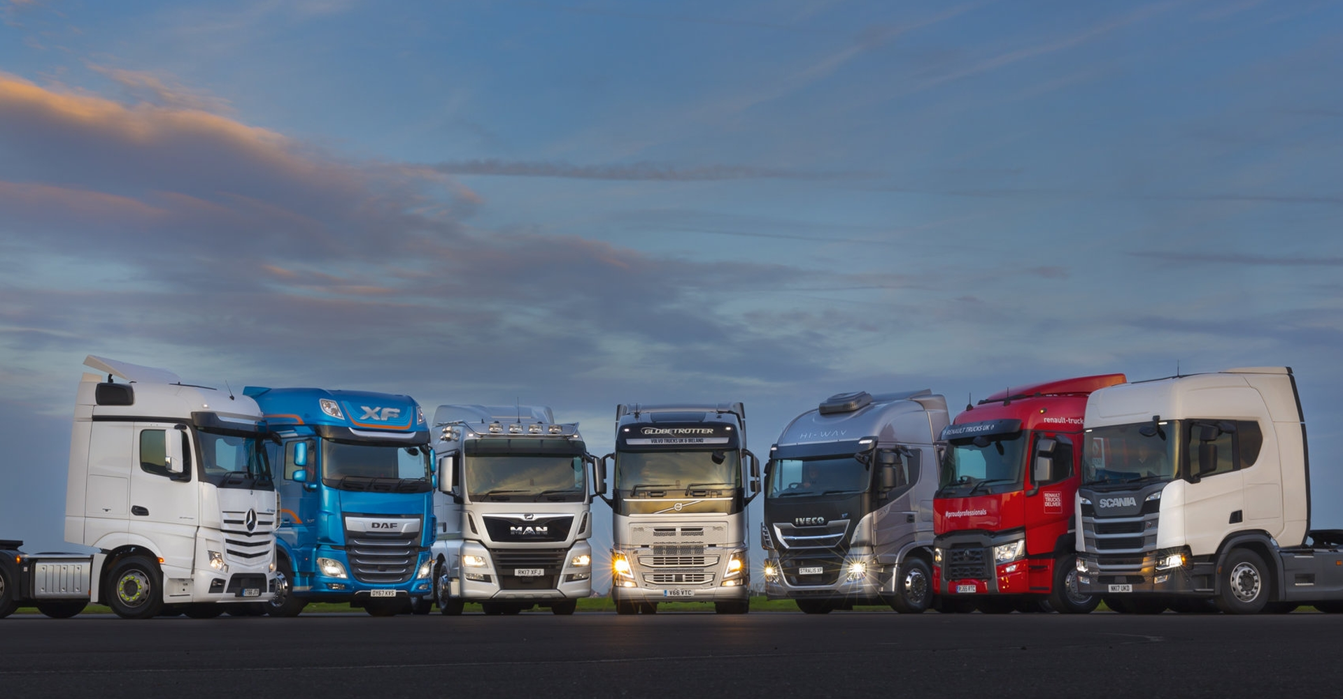 Seven Inspire E-SES lorries lined up in a slight curvature, with a cloudy sky behind them