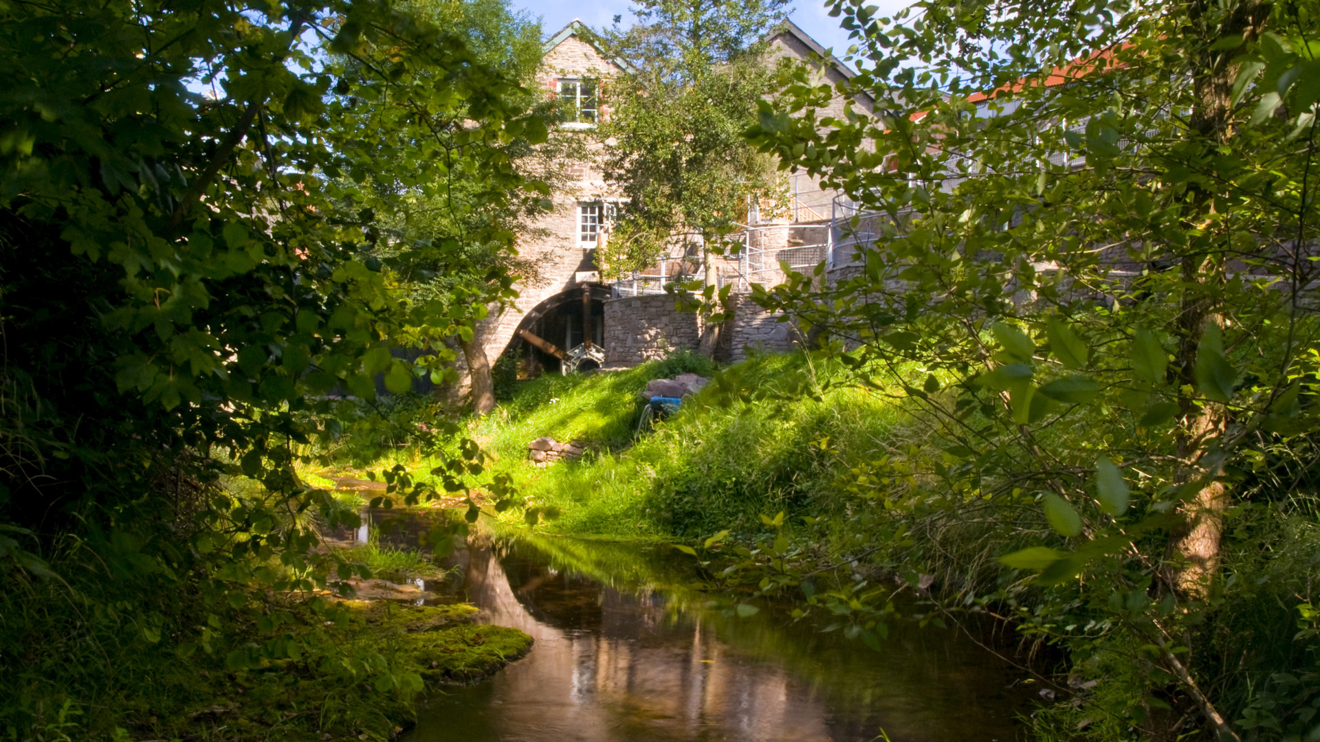 A beautiful view of Talgarth Mill. Surrounded by green tree's and grass banks, with a small river running through the centre.