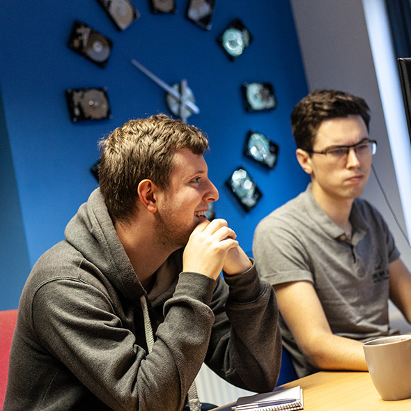 two web developers looking focused in office room with large clock behind them
