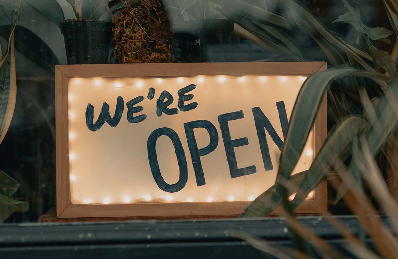 A lit up wooden frame with 'We're Open' written on it, hidden between some plants in a window