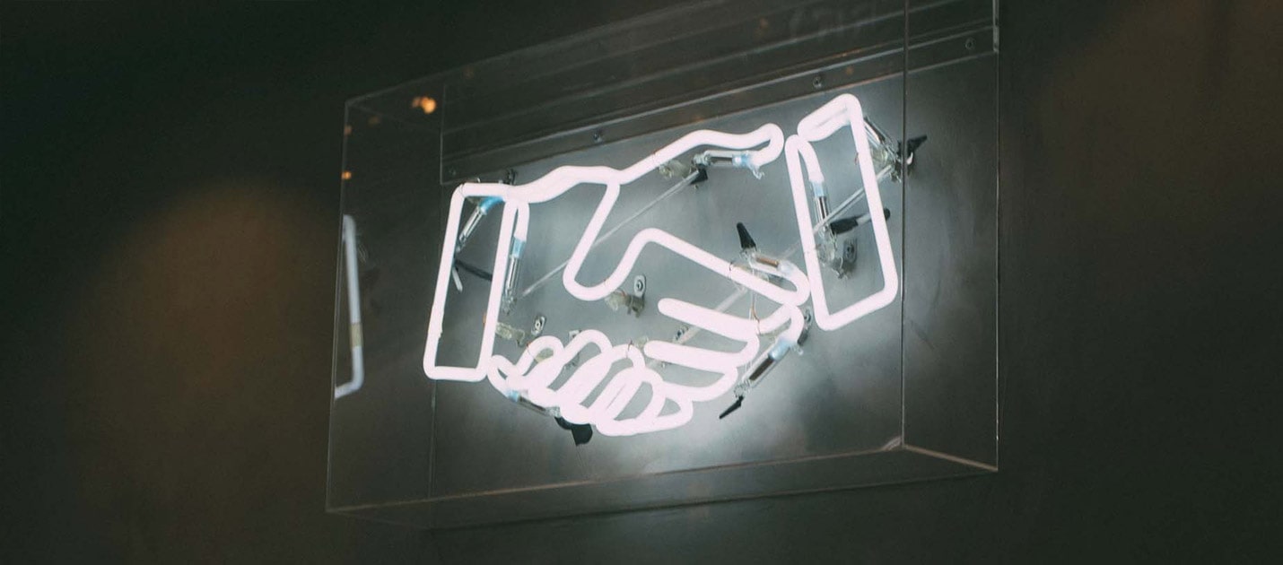 A LED white sign showing two hands shaking hands in a clear casing on a grey wall