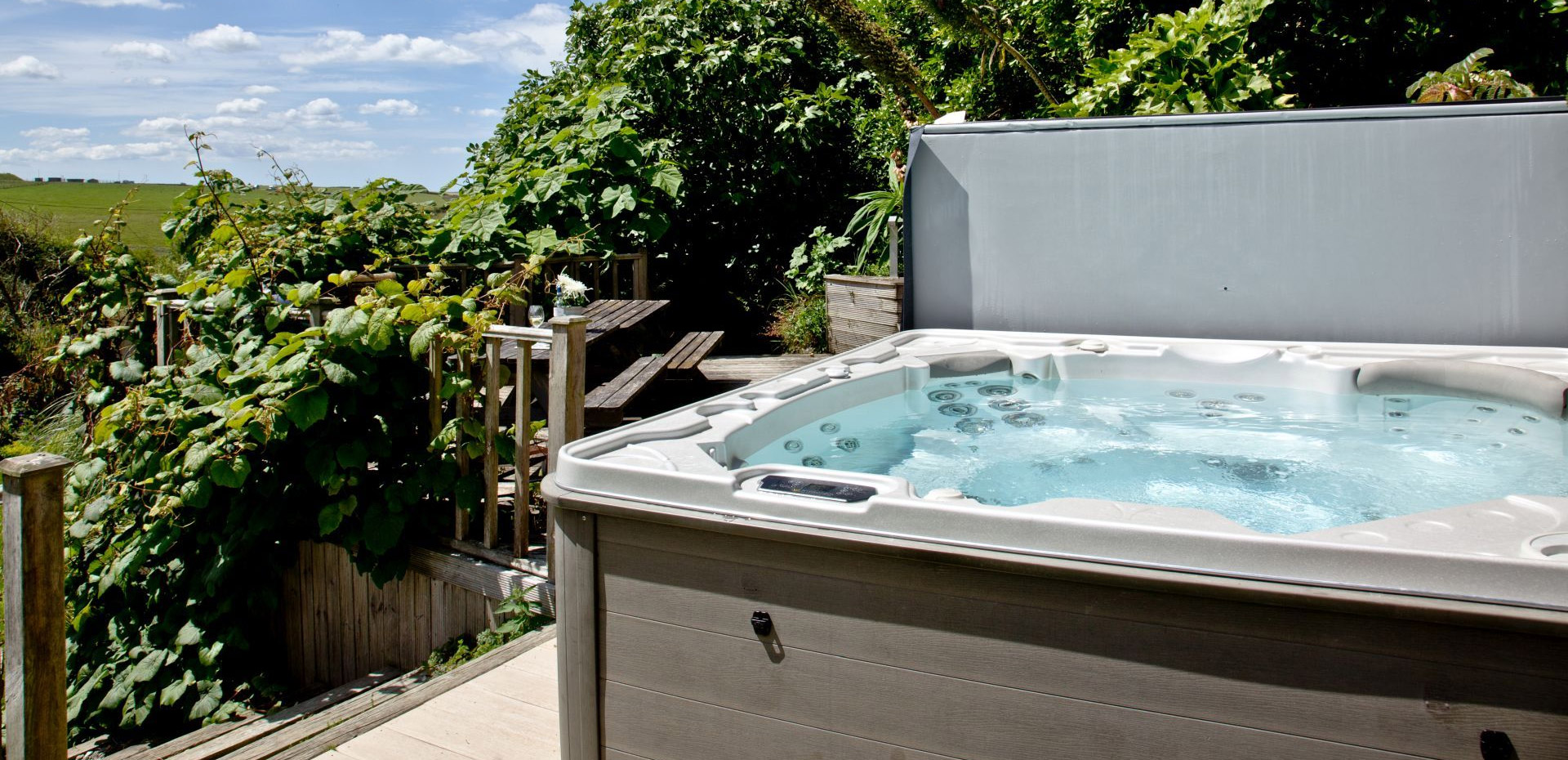 An open hot tub surronded by greenery, wooden decking and blue skies